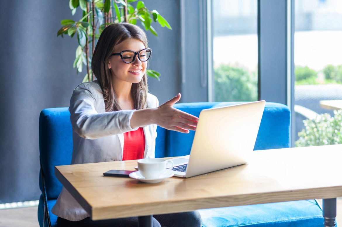 Portrait of happy beautiful stylish young woman in glasses sitting, looking at her laptop screen on video call and giving hand to handshake, toothy smile.
